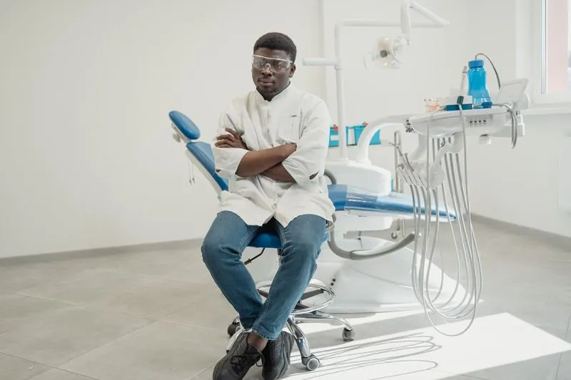 A dentist in a white coat seated in a treatment room with his arms folded